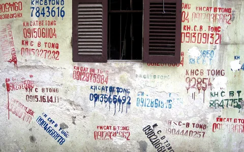 Verwitterte Hauswand mit einem geschlossenen braunen Fenster in der Mitte, dessen Läden offen stehen. Die Wand ist mit roten, blauen und schwarzen Telefonnummern und dem Schriftzug KH CẮT BÊ TÔNG besprüht.