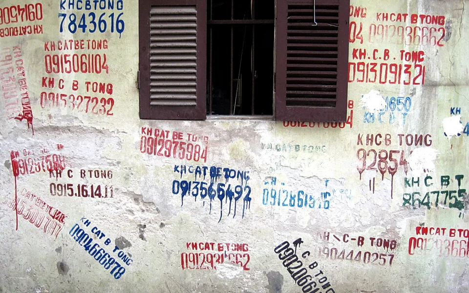 Verwitterte Hauswand mit einem geschlossenen braunen Fenster in der Mitte, dessen Läden offen stehen. Die Wand ist mit roten, blauen und schwarzen Telefonnummern und dem Schriftzug KH CẮT BÊ TÔNG besprüht.