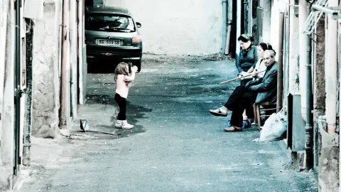 Kleines Mädchen steht auf einer schmalen Gasse, rechts sitzen drei Erwachsene auf Stühlen, links parkt ein dunkles Auto.