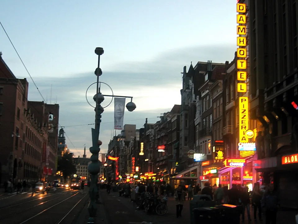 Belebte Straße in Amsterdam in der Dämmerung mit gelben und roten Leuchtreklamen, darunter das Damhotel und eine Pizzeria, links eine Straßenbahnschiene und Autos.