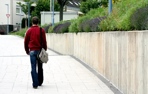 Ein Mann in roter Jacke und Jeans geht von hinten gesehen einen gepflasterten Gehweg entlang, er trägt eine graue Umhängetasche. Rechts verläuft eine helle Betonmauer, darüber wachsen Lavendel und Gräser.