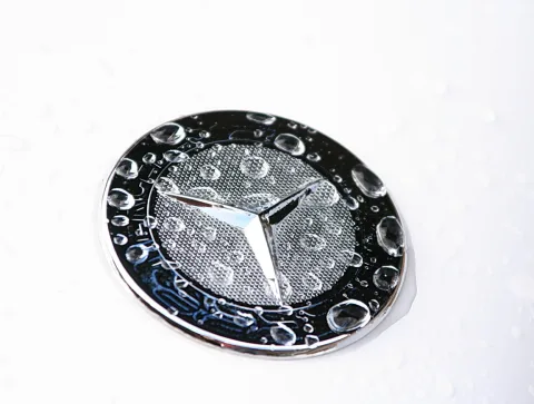 Chromfarbener Mercedes-Stern auf einem runden Emblem, übersät mit Wassertropfen, vor weißem Untergrund.