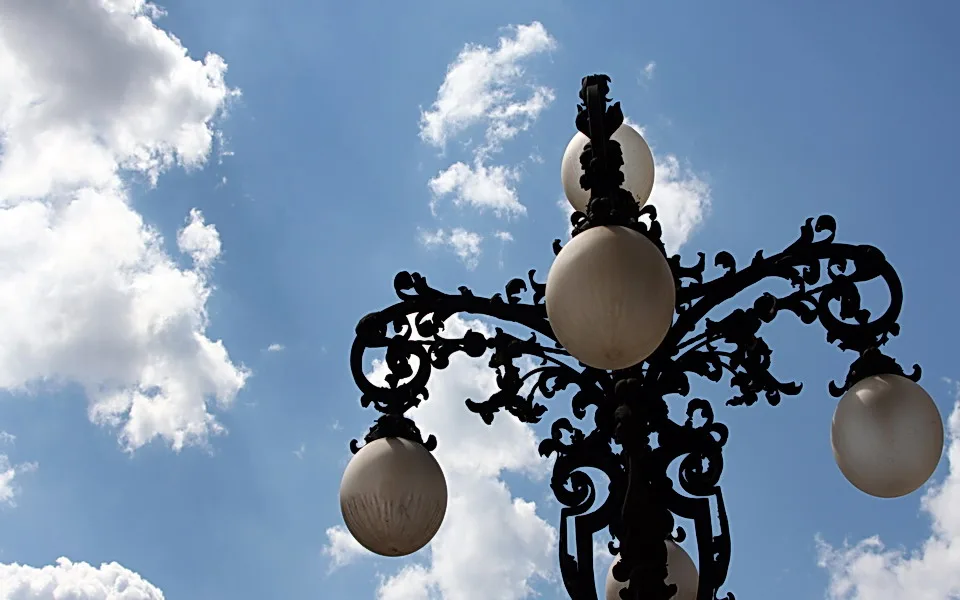 Schwarze, reich verzierte schmiedeeiserne Laterne mit vier milchweißen Glaskugeln vor blauem Himmel mit weißen Wolken, von unten fotografiert.
