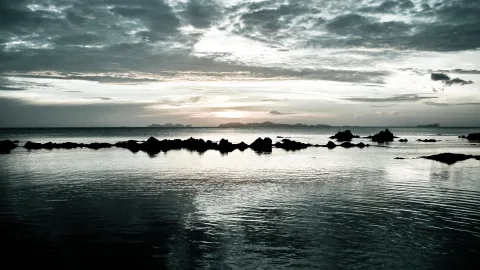Dunkle Felsen ragen aus ruhigem Meer, darüber dichte Wolken und eine tief stehende Sonne am Horizont.