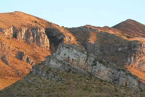 Steile Felswände mit hellen Gesteinsschichten an einem Berghang, von tief stehender Abendsonne orange angestrahlt, darüber ein klarer blassblauer Himmel.