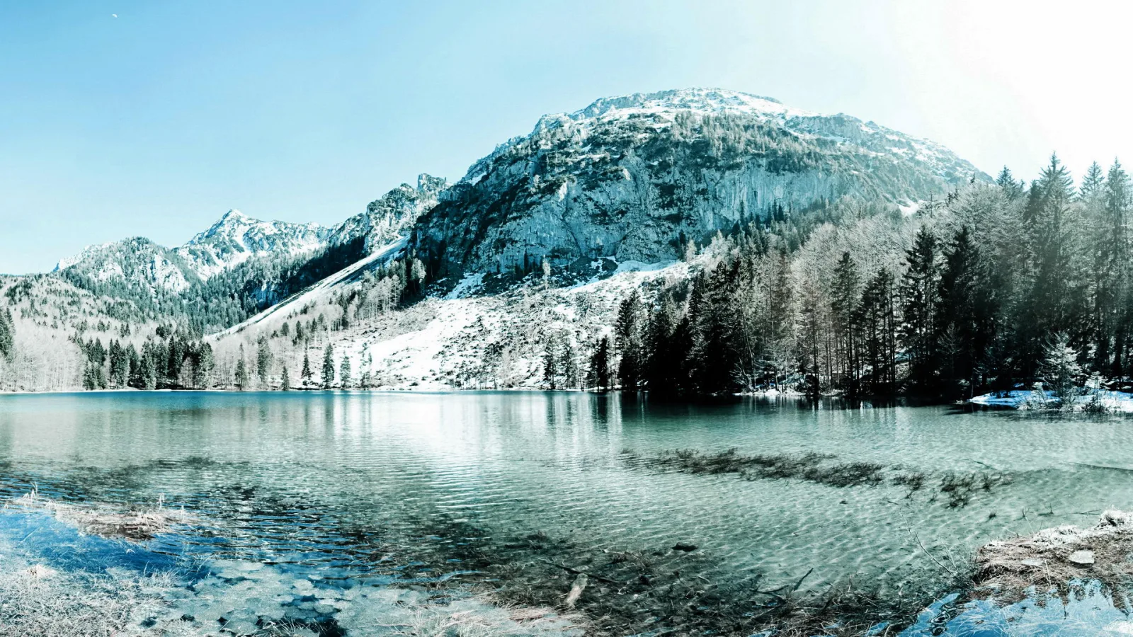 Ruhiger Bergsee mit türkisfarbenem Wasser, im Hintergrund steile, teils verschneite Felswände und dichter Nadelwald unter klarem blauen Himmel.