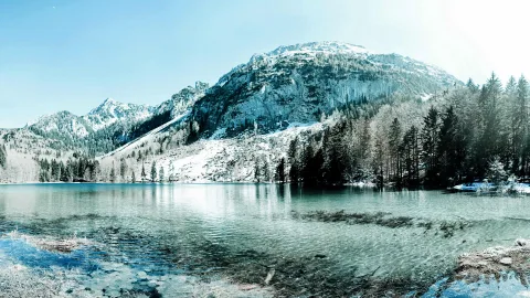 Ruhiger Bergsee mit türkisfarbenem Wasser, im Hintergrund steile, teils verschneite Felswände und dichter Nadelwald unter klarem blauen Himmel.