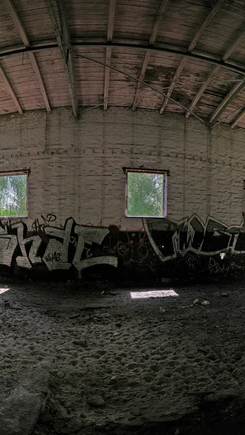 Leere, verlassene Halle mit gewölbter Holzdecke, weiß gekalkter Backsteinwand, zwei Fenstern mit Blick auf Bäume und Graffiti an der unteren Wand, sandiger Boden.