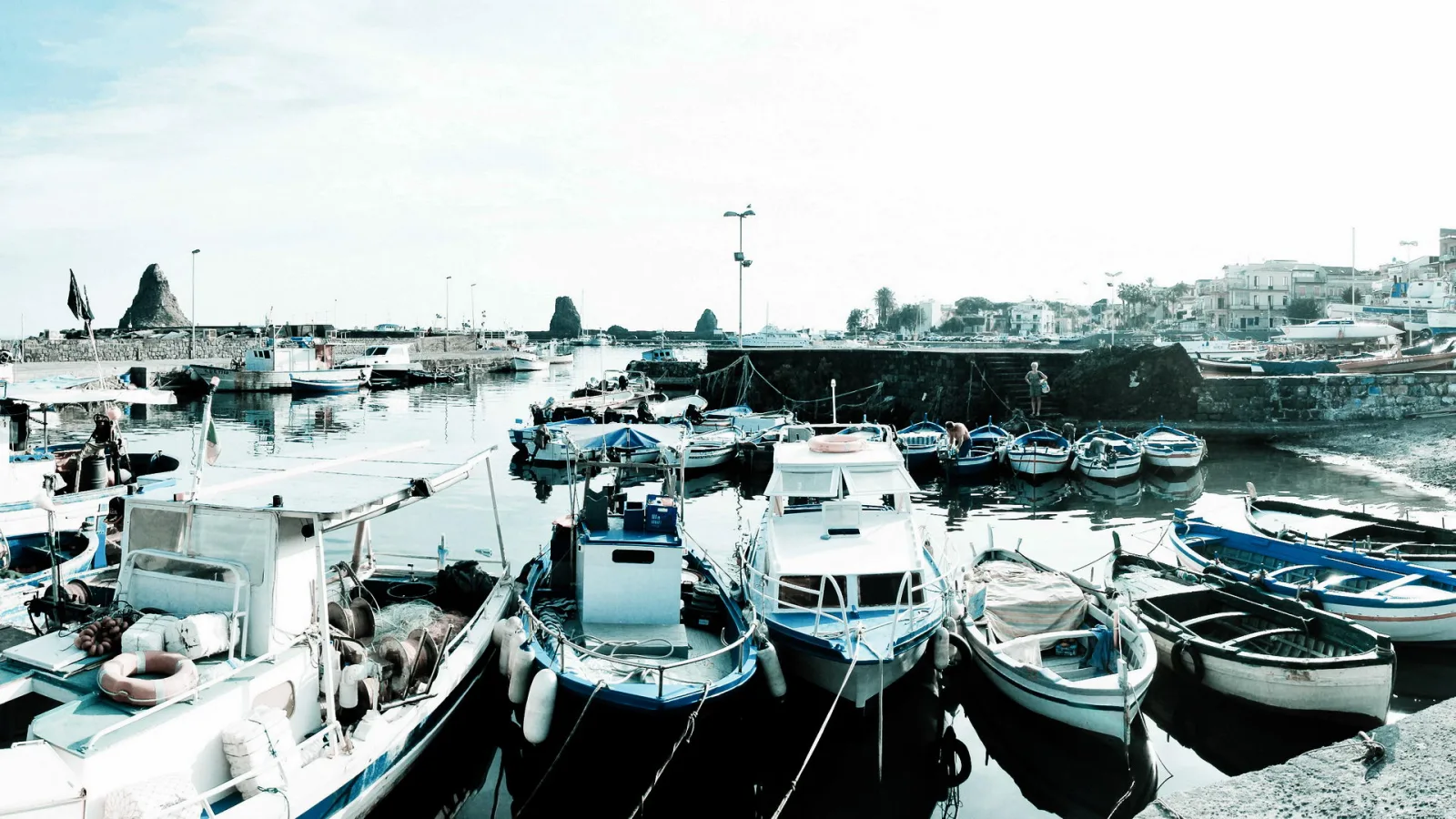 Mehrere kleine Boote mit blauen und weißen Rümpfen liegen im ruhigen Hafenbecken vor einer Mole, im Hintergrund stehen Häuser und zwei dunkle Felsen im Meer unter hellem Himmel.