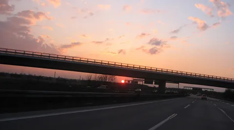 Autobahn unter einer Brücke bei Sonnenuntergang, die rote Sonne steht tief am Horizont, Autos fahren auf der Fahrbahn, der Himmel ist rosa und orange mit Wolken.