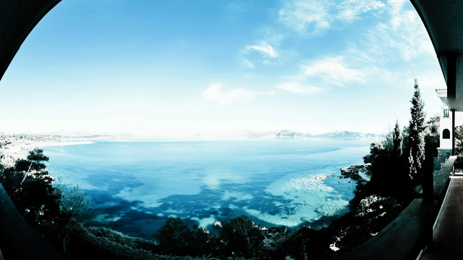 Breites Panorama auf eine blaue Meeresbucht mit Kiefern im Vordergrund, einer Villenterrasse rechts und einer Küstenlinie mit Bergen am Horizont.