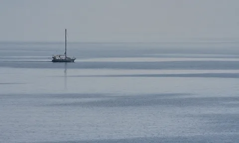 Ein einzelnes weißes Segelboot mit hohem Mast fährt auf einer glatten, grau-blauen Meeresfläche unter bedecktem, dunstigem Himmel.