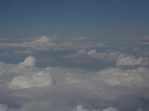 Blick aus großer Höhe auf eine dichte Wolkendecke mit weichen, weißen Haufenwolken unter einem blassblauen Himmel.