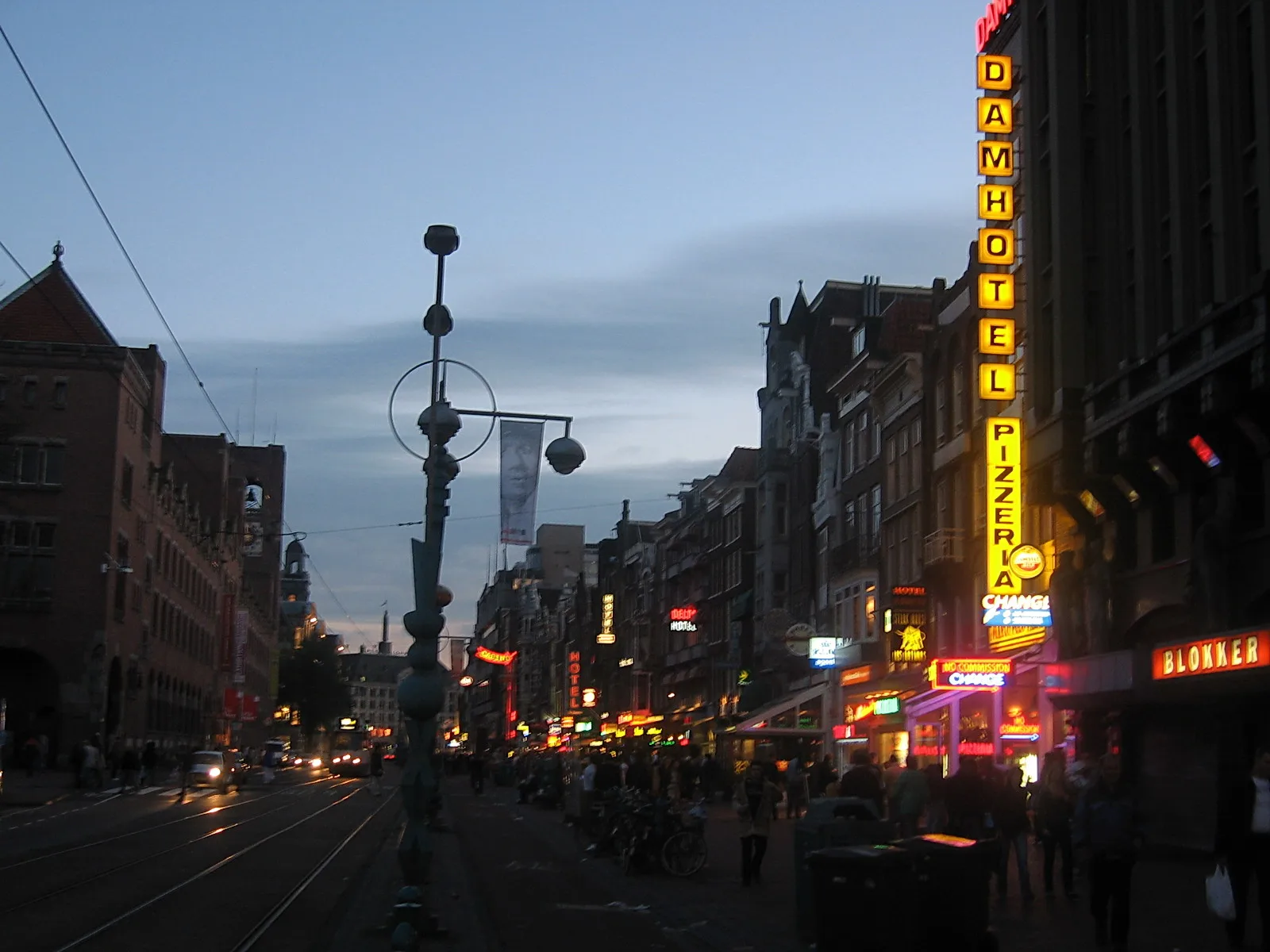 Abendliche Straße in Amsterdam mit einer hohen vertikalen Leuchtreklame für das Damhotel und einer Pizzeria, daneben Straßenbahngleise, Laternen, Fahrräder und Passanten.