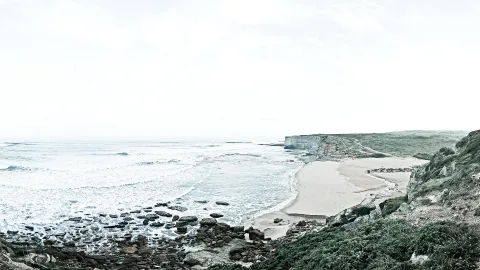 Sandstrand und Felsen an einer Küste: Links brechen Wellen auf dunkle Steine, rechts steigt grün bewachsenes Kliff an, darüber liegt heller, bewölkter Himmel.