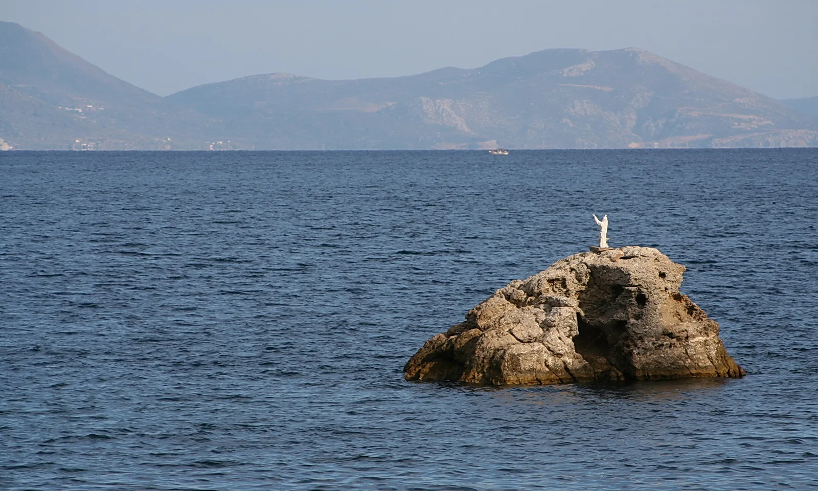 Ein einzelner Felsen ragt aus ruhigem blauem Meer, obenauf steht eine weiße Figur mit erhobenen Armen. Dahinter liegt eine dunstige Gebirgskette.