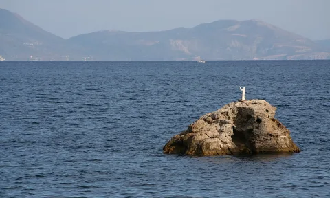 Ein einzelner Felsen ragt aus ruhigem blauem Meer, obenauf steht eine weiße Figur mit erhobenen Armen. Dahinter liegt eine dunstige Gebirgskette.