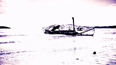 Ein halb versunkenes, verrostetes Holzboot ragt aus flachem Wasser, daneben eine dunkle Stange, im Hintergrund schmale Küstenstreifen unter einem hellen, lila getönten Himmel.