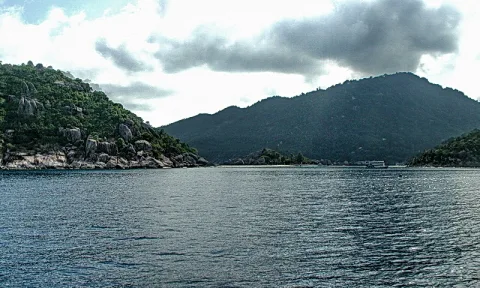 Blick aufs offene Meer mit dunkelgrünen bewaldeten Hügeln links und rechts, im Vordergrund felsige Küste, darüber bewölkter Himmel