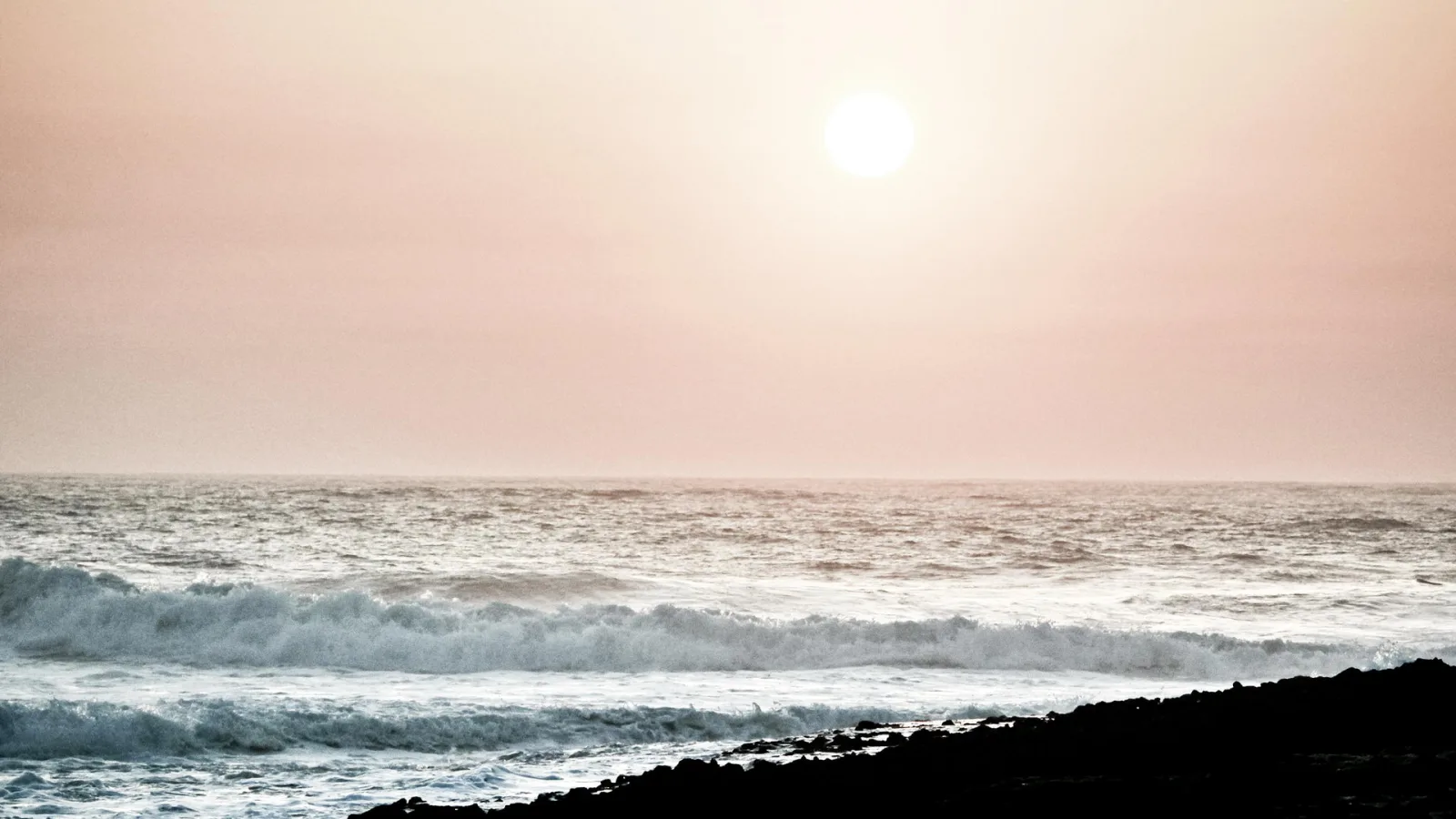 Weiße Wellen brechen an einem dunklen Steinstrand, darüber steht eine blasse Sonne im rosafarbenen Abendhimmel über einem ruhigen Meer.