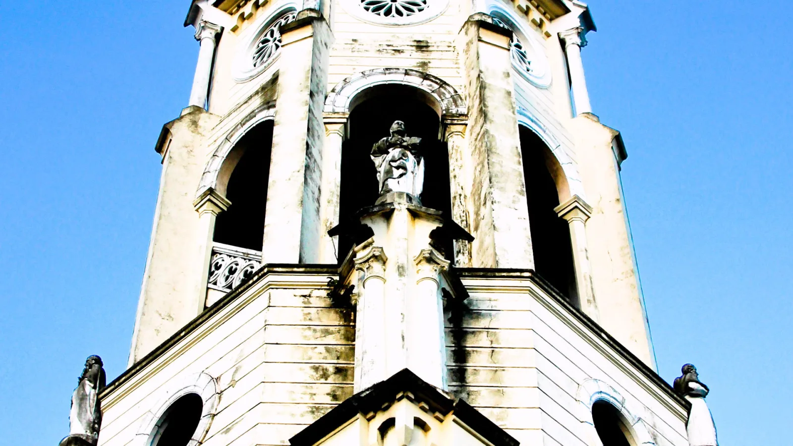Heller Kirchturm mit Statuen in Nischen und Eckfiguren, von unten vor blauem Himmel fotografiert.