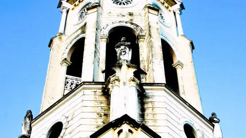 Heller Kirchturm mit Statuen in Nischen und Eckfiguren, von unten vor blauem Himmel fotografiert.