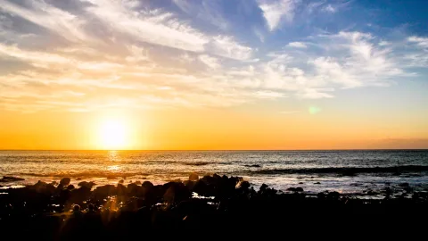 Sonnenuntergang über dem Meer, die Sonne steht tief am Horizont und spiegelt sich auf dem Wasser. Im Vordergrund dunkle Felsen am Ufer, darüber ein Himmel mit Schleierwolken in Orange, Gelb und Blau.