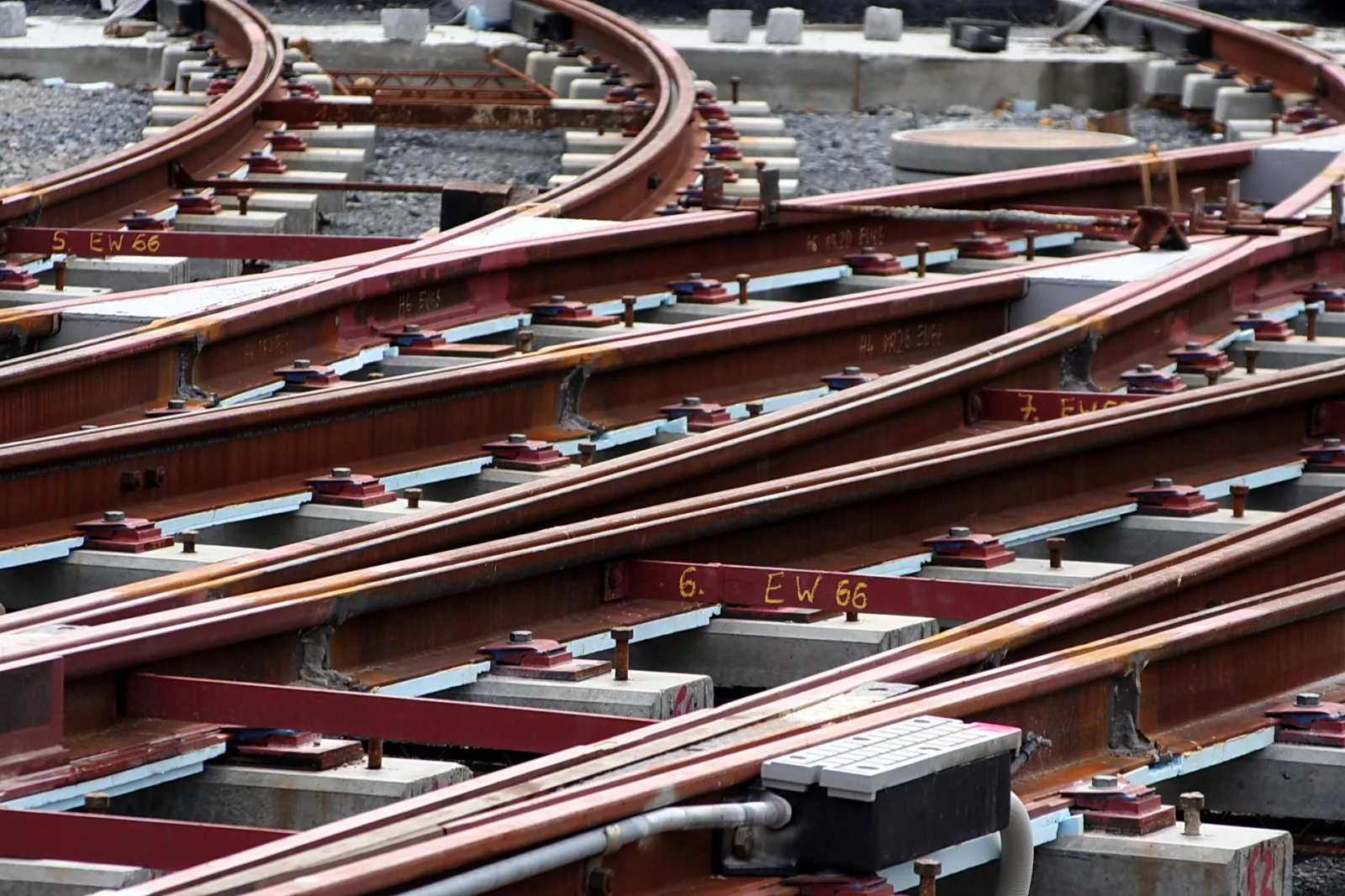 Mehrere rostrote Schienen einer Gleisweiche auf grauen Betonschwellen, mit Schraubenbefestigungen und gelber Markierung "6. EW 66" auf dem Schienenstrang.