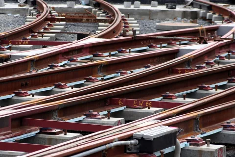Mehrere rostrote Schienen einer Gleisweiche auf grauen Betonschwellen, mit Schraubenbefestigungen und gelber Markierung "6. EW 66" auf dem Schienenstrang.