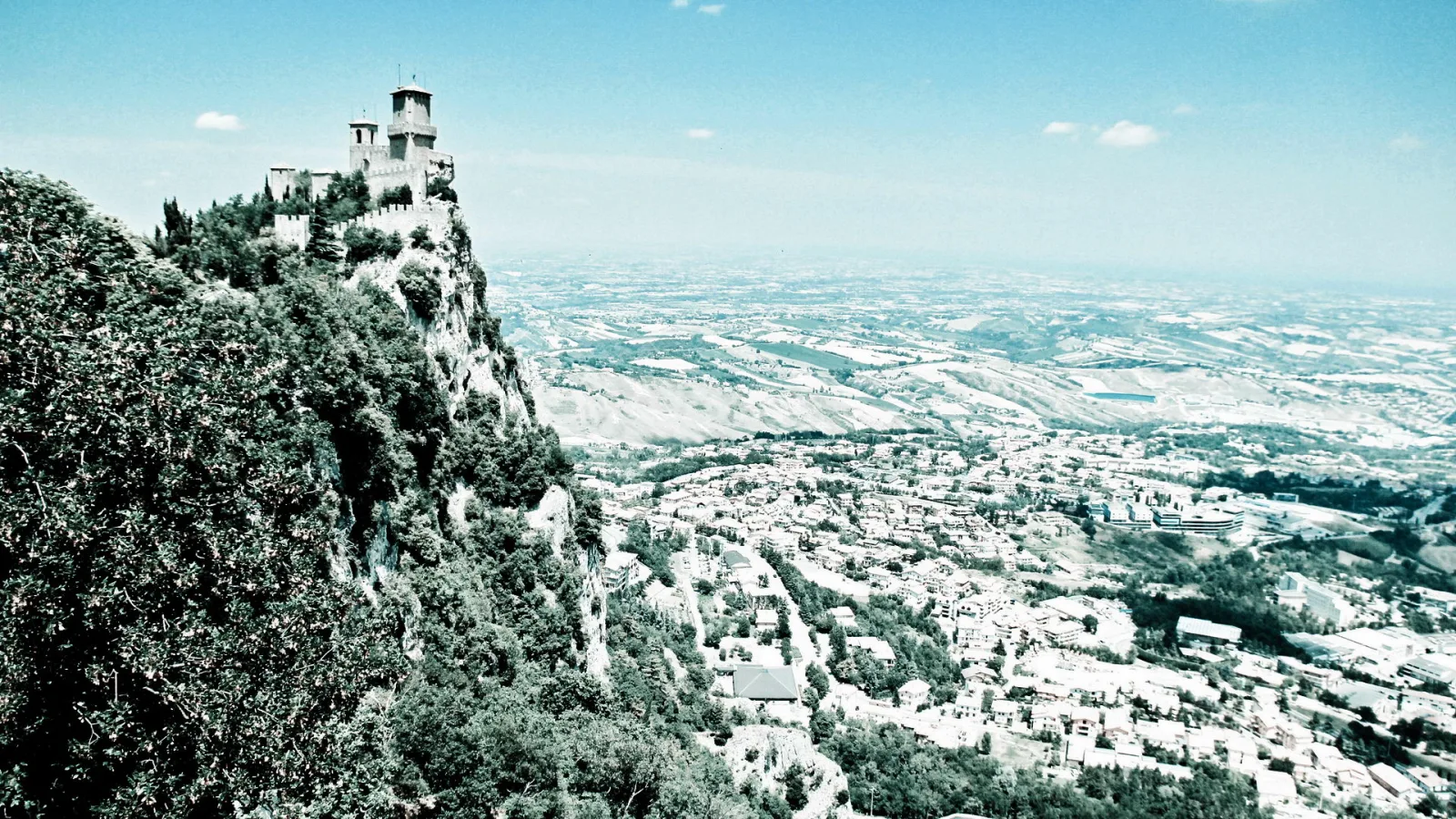 Eine alte Steinburg mit Türmen steht auf einem bewaldeten Felsvorsprung, darunter liegt ein Dorf mit dicht stehenden Häusern und weites Hügelland unter hellblauem, dunstigem Himmel.