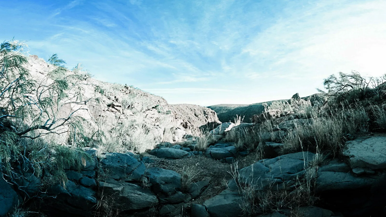 Felsige Schlucht mit grauen Felsbrocken im Vordergrund, trockenem Gestrüpp und hellen Felswänden unter blauem Himmel mit feinen Wolkenschleiern.