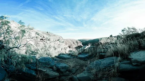 Felsige Schlucht mit grauen Felsbrocken im Vordergrund, trockenem Gestrüpp und hellen Felswänden unter blauem Himmel mit feinen Wolkenschleiern.