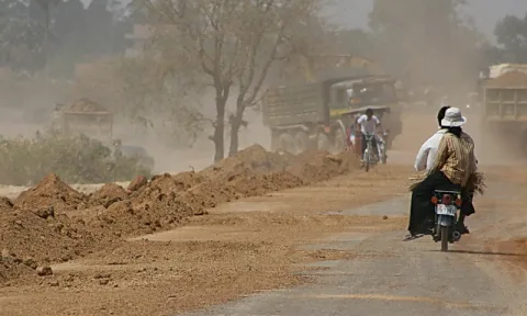 Staubige unbefestigte Straße in Phnom Penh mit aufgeschütteten Erdhaufen links, einem Mofa mit zwei Personen im Vordergrund und mehreren Lastwagen im Staub dahinter.