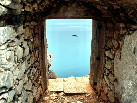 Durchgang in einer Steinmauer, der den Blick auf ruhiges blaues Meer freigibt. Ein kleines Schiff fährt am Horizont.