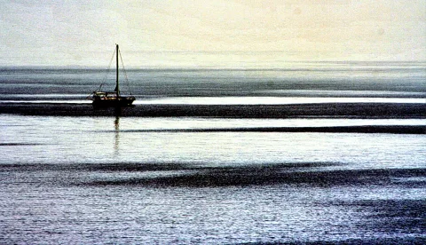 Ein einzelnes Segelboot auf ruhigem, grauem Wasser mit flachen Sandbänken unter diesigem Himmel.