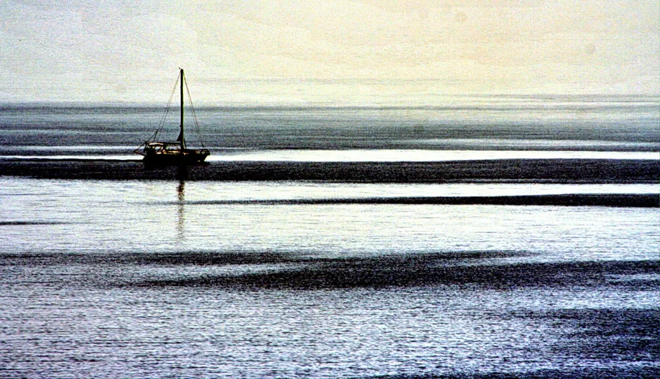 Ein einzelnes Segelboot auf ruhigem, grauem Wasser mit flachen Sandbänken unter diesigem Himmel.