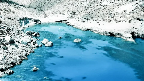 Türkisblaues Wasser einer kleinen Bucht, am linken Ufer liegen Felsbrocken, rechts und oben ziehen sich helle, karge Hänge hin.