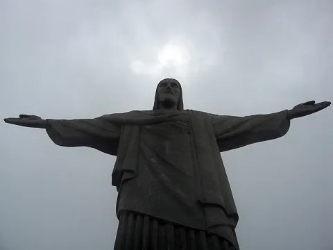 Große Christusstatue mit weit ausgebreiteten Armen vor grauem, bewölktem Himmel, von unten fotografiert.