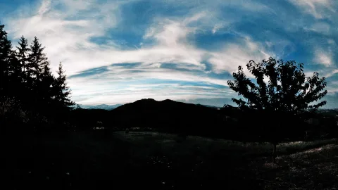 Panoramablick auf einen dunklen Hügel am Abend, links Nadelbäume, rechts ein Baum vor blauem Himmel mit Schleierwolken.