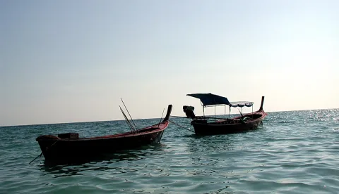 Zwei hölzerne Langschwanzboote liegen auf ruhigem, blaugrünem Meer, dahinter ein heller, dunstiger Himmel.