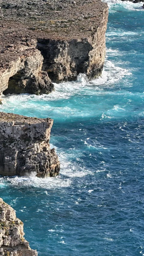 Türkisblaues Meer schlägt mit weißer Gischt gegen zerklüftete Felsklippen, links oben und links unten ragen helle Steinkanten ins Wasser.