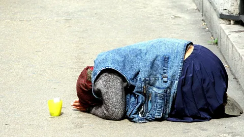 Eine Person kniet mit dem Gesicht zum Boden auf grauem Asphalt, daneben steht ein gelber Becher. Sie trägt eine Jeansjacke und einen dunkelblauen Rock.
