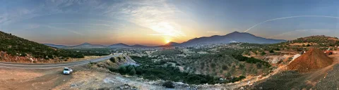 Panoramablick bei Sonnenuntergang auf ein weites Tal mit Olivenhainen, einer Stadt am Fuß eines Gebirges und einer kurvigen Straße im Vordergrund, dazu ein weißes Auto und ein rötlicher Erdhaufen rechts.