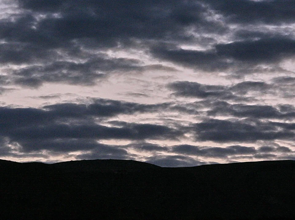 Dunkle Hügelkette im unteren Bildbereich, darüber ein Band aus grauen und bläulichen Wolken am Abendhimmel