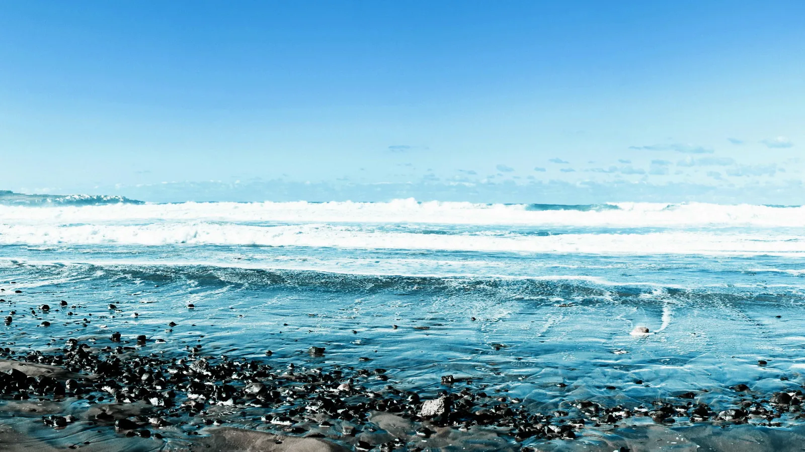 Weiße Brandung rollt über blaues Meer, im Vordergrund liegen dunkle Kiesel am Ufer unter klarem blauem Himmel.