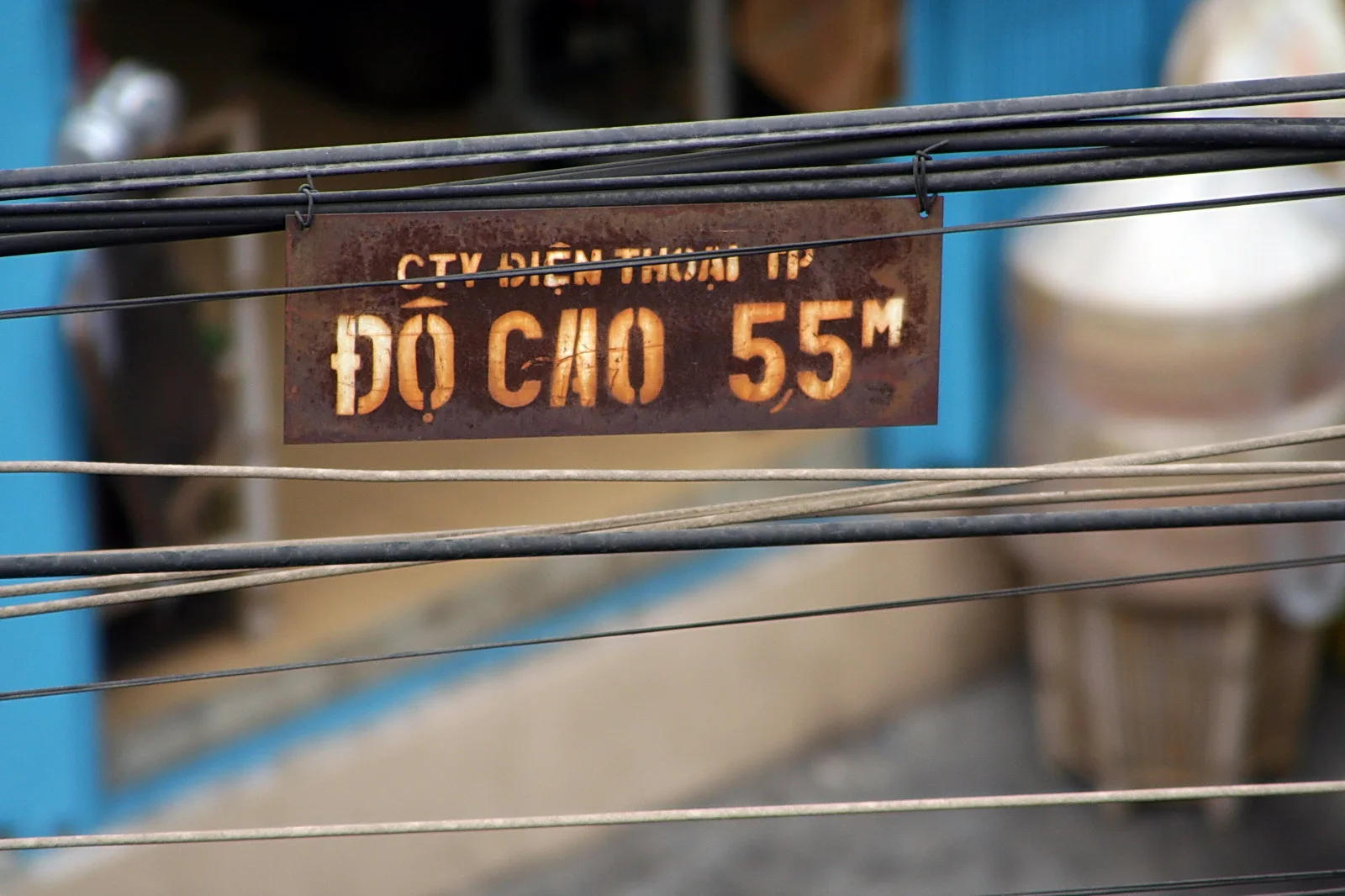 Verrostetes Metallschild mit vietnamesischer Aufschrift „Độ cao 55m“ hängt zwischen schwarzen Stromkabeln vor einer blau gestrichenen Wand.