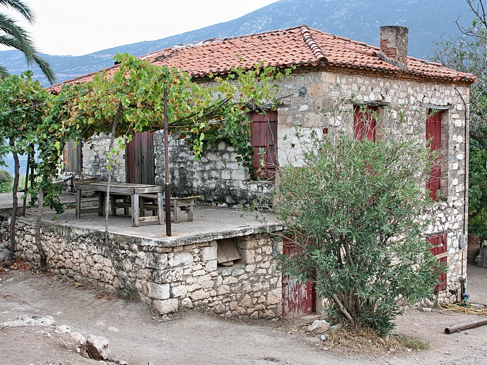 Altes Steinhaus mit rotem Ziegeldach, überwuchert von einer Weinlaube, davor ein Holztisch und ein Oleanderstrauch, im Hintergrund Berge.