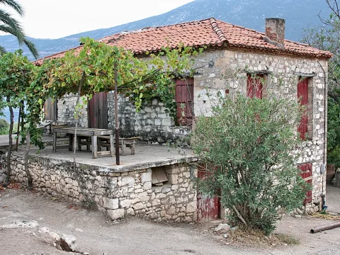 Altes Steinhaus mit rotem Ziegeldach, überwuchert von einer Weinlaube, davor ein Holztisch und ein Oleanderstrauch, im Hintergrund Berge.