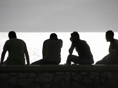 Vier Männer sitzen im Gegenlicht auf einer Steinmauer, vor einer hellen Wasserfläche und einem grauen Himmel.