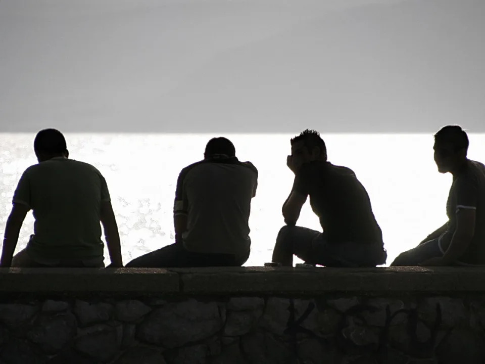 Vier Männer sitzen im Gegenlicht auf einer Steinmauer, vor einer hellen Wasserfläche und einem grauen Himmel.